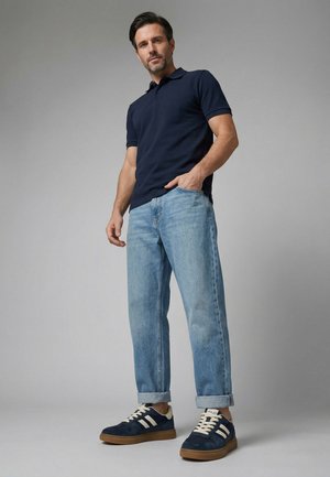 Mand stående med den ene hånd i jeanslommen, iført en marineblå polo-shirt, lyseblå op rullede jeans og marineblå sneakers med hvide striber.