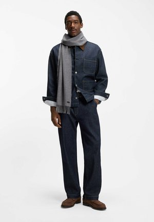 Kurtka dżinsowa na guziki, z dwoma przednimi kieszeniami i brązowym kołnierzem, zestawiona z ciemnymi dżinsowymi spodniami i jasnoszarym szalikiem. Całość dopełniają brązowe buty.