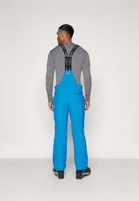 Grenouillère bleue avec des bretelles noires ajustables et une texture lisse. Stylisée avec une poche arrière et ajustée au niveau des jambes.