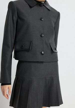 Veste cintrée gris foncé avec boutons noirs et rabats de poche associée à une jupe plissée assortie portée par une personne debout.