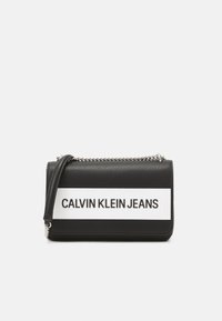Černá texturovaná kabelka s bílým horizontálním pruhem, na kterém je tučně vytištěno "CALVIN KLEIN JEANS". Obsahuje řetízkový popruh.