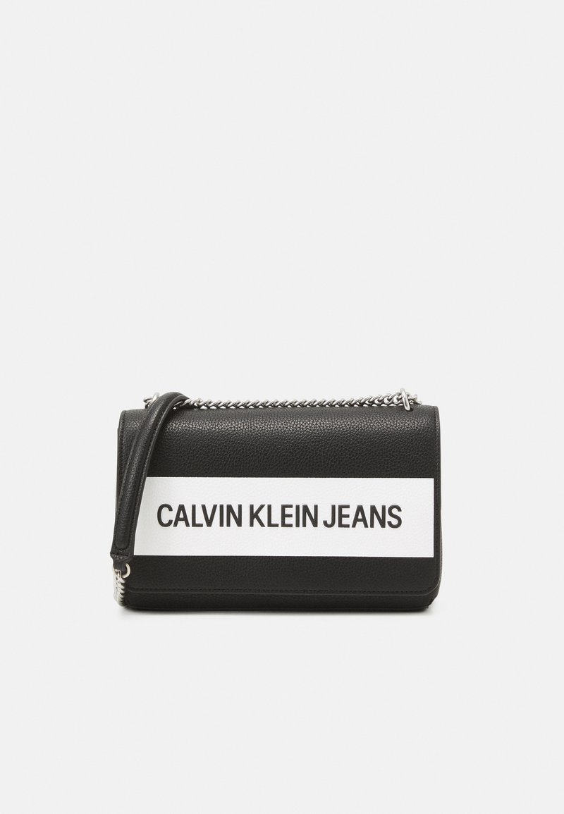 Černá texturovaná kabelka s bílým horizontálním pruhem, na kterém je tučně vytištěno "CALVIN KLEIN JEANS". Obsahuje řetízkový popruh.