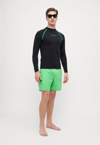 Schwarzes Langarm-Rashguard mit grünen Akzenten und Logo, kombiniert mit leuchtend grünen Badehosen für einen abgestimmten Badelook.