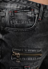 Black denim jeans featuring a zippered pocket and contrasting red stitching. The fabric has a faded texture and a fitted design.