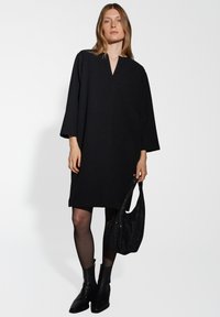 Zwart oversized jurk met een V-hals, lange mouwen, met een gestructureerde stof. Model houdt een zwart paillettentas vast en draagt glanzende enkellaarsjes.
