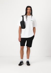 Camiseta blanca de gran tamaño con texto verde, pantalones cortos negros, bolso negro de hombro y zapatillas negras con rayas blancas. Fondo liso.