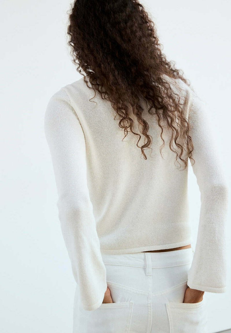 Personne avec de longs cheveux bouclés portant un haut blanc à manches longues et un pantalon blanc, les mains dans les poches arrière, dos au spectateur.