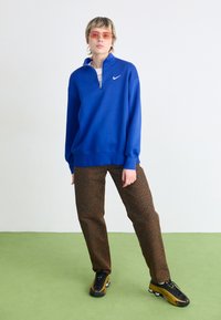 Sudadera azul con cremallera y mangas largas, combinada con pantalones marrón con estampado de leopardo y zapatillas negras con amarillo. Modelo posa sobre un suelo verde.