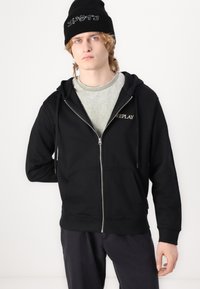 Felpa nera con zip, tasca frontale e cappuccio con cordino, con il logo "REPLAY" in oro. Indossata sopra una maglietta grigia chiara e abbinata a pantaloni scuri.