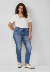 Kobieta z kręconymi włosami stoi przy szarej ścianie, ubrana w białą koszulkę z dekoltem w serek, niebieskie dżinsy i beżowe sneakersy.