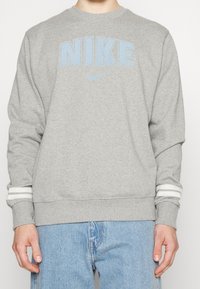 Šedá mikina s bledě modrým logem "NIKE". Má bílé pruhované manžety. Vyrobena z měkké, texturované tkaniny. Kulatý výstřih a volný střih.