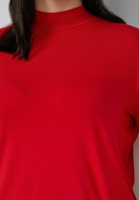 Czerwona bluzka z długim rękawem i wysokim kołnierzem, wykonana z gładkiego materiału. Dopasowany krój z minimalną ilością szwów i jednolitym kolorem.