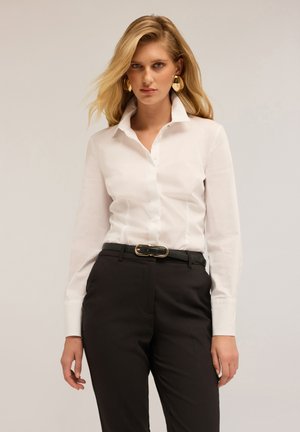 Chemise blanche à col structuré, associée à un pantalon noir ajusté et une ceinture noire texturée avec une boucle dorée.