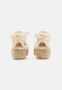 Tan espadrille heels with a jute rope platform, open back, and cream fabric ankle strap tied in bows. Textured surface, casual design.