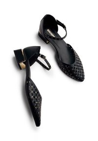 Zapatos planos negros con puntera puntiaguda, patrón geométrico recortado, tira en el tobillo y acento dorado en el tacón. Textura de cuero suave.