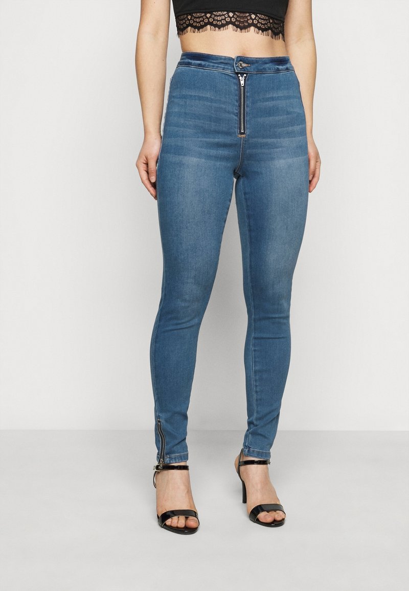 Pantalones vaqueros ajustados de mezclilla azul con cintura alta, cremallera frontal y detalles de cremallera en los puños. Textura suave con un ligero desgaste.