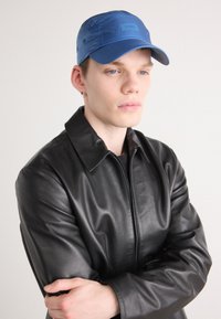 Black leather jacket with a zip front and wide collar, paired with a blue cap featuring a textured surface and logo patch.