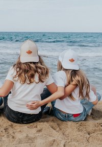 Zwei Personen sitzen an einem Sandstrand, tragen weiße und beige Kappen mit Logos. Sie haben lange Haare und lässige weiße T-Shirts und Jeans.