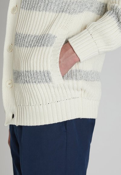 Cárdigan de punto en color crema con rayas grises, textura acanalada, cierre de botones y bolsillos laterales. Combinado con pantalones azul oscuro.