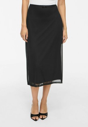 Femme portant une jupe midi noire avec des panneaux latéraux transparents, associée à des talons ouverts noirs et un haut blanc, debout devant un fond blanc.