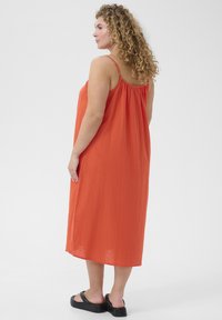 Orange sleeveless dress with a flowing design and a gathered back. Model wears black platform sandals, with a textured fabric finish.