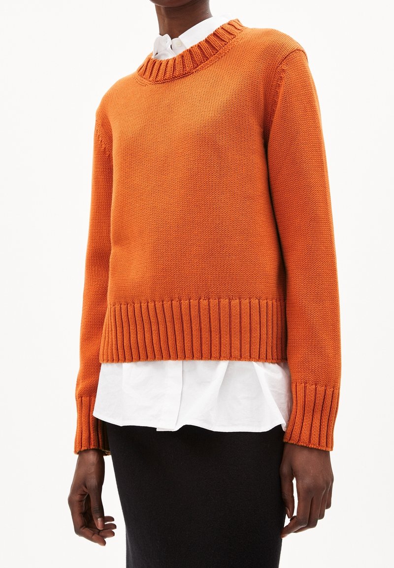 Personne portant un pull en tricot orange par-dessus une chemise blanche, associé à une jupe noire, debout sur un fond blanc.