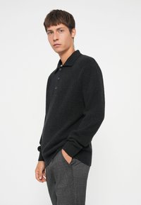 Schwarzer Strickpullover mit Kragen und Knopfleiste mit drei Knöpfen; strukturierter Stoff. Getragen mit grauen karierten Hosen, Hände in den Taschen.