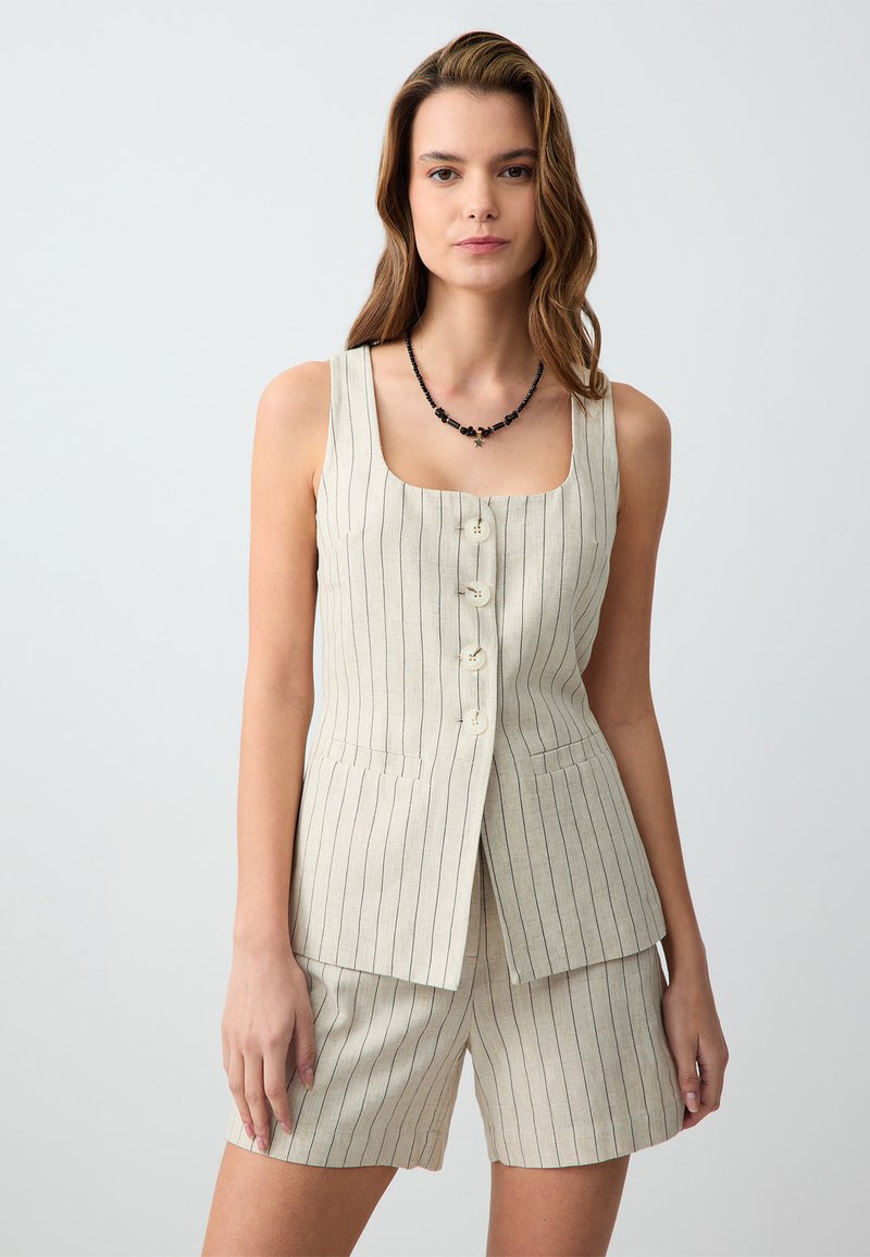 Gilet in lino beige con righe verticali, scollatura quadrata e chiusura frontale a cinque bottoni. Abbinato a pantaloni sartoriali coordinati e una collana di perline nere.