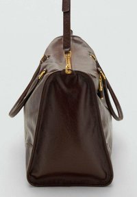 Sac de sport en cuir marron foncé avec des ferrures dorées et une bandoulière réglable, présenté de l'extrémité étroite sur un fond uni.