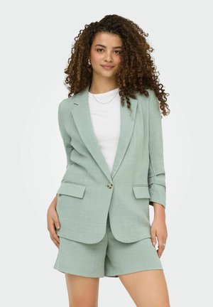 Donna con capelli ricci che indossa un blazer verde menta e pantaloncini coordinati sopra una camicia bianca, in piedi contro uno sfondo bianco.