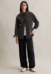 Ciemnoszara koszula sztruksowa z długim rękawem, z przednią kieszenią, noszona na białej koszuli, w połączeniu z luźnymi czarnymi spodniami oraz kremowymi sneakersami.