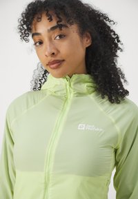 Giacca leggera verde chiaro con zip di Jack Wolfskin, caratterizzata da una texture morbida e liscia, colletto alto e dettaglio del logo sul lato sinistro del petto.