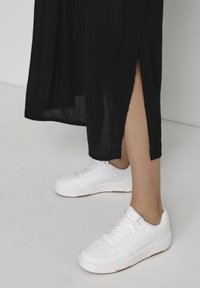 Sneaker sportive bianche in pelle leggermente texturizzata, con suola piatta in gomma e chiusura con lacci, abbinate a un vestito nero con uno spacco laterale.