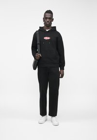 Μαύρο φούτερ με κουκούλα και κόκκινο λογότυπο στο στήθος, συνδυασμένο με μαύρο παντελόνι και λευκά αθλητικά παπούτσια. Το μοντέλο κρατάει μια μαύρη τσάντα ώμου.