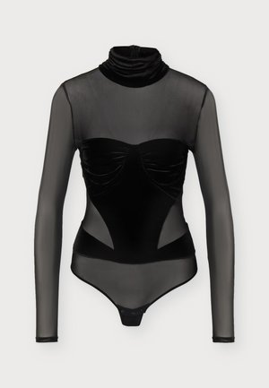 Un mannequin se tient devant un fond clair uni, vêtu d’un haut noir transparent à manches longues, d’un pantalon à jambes larges et de chaussures pointues.