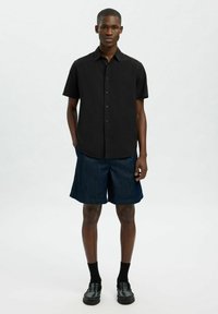 Camisa negra de manga corta con botones combinada con pantalones cortos de mezclilla azul oscuro, calcetines negros y zapatos negros con un acabado texturizado.