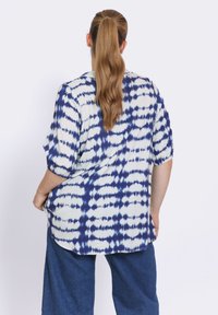 Korte mouwen shirt in het wit met een blauw tie-dye patroon, met een losse pasvorm en een gebogen zoom. Gepaarde met donkerblauwe spijkerbroek.