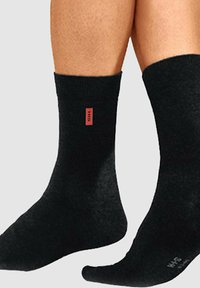 Schwarze Socken aus einem strukturierten Stoff, mit einem roten Logoetikett auf einer Seite. Knöchelhohes Design mit einem glatten, einheitlichen Erscheinungsbild.