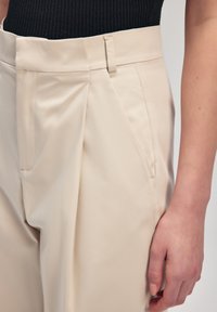 Pantaloni beige a vita alta realizzati in tessuto liscio, con pieghe, una tasca frontale e un design a gamba larga.