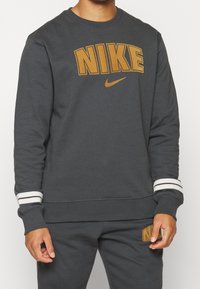 Sudadera gris oscuro con un gran logotipo naranja de "NIKE" y rayas de acento en las mangas. Presenta puños acanalados y una textura suave.