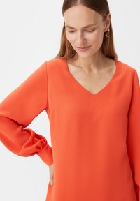Kleid mit V-Ausschnitt und langen Ärmeln aus leuchtend orangefarbenem Stoff mit glatter Textur und sanft gepufften Ärmeln. Verfügt über eine dezente Seitentasche.
