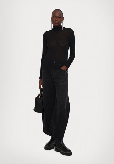 Top negru din material transparent cu maneci lungi și guler înalt, asortat cu blugi negri cu talie înaltă și croi larg. Modelul ține în mână o mică geantă neagră, purtând ghete negre masive.