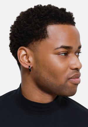 Zwarte ronde hoepeloorketting met een matte afwerking, weergegeven op een model dat een zwart coltrui draagt en kort, textuurhaar heeft.