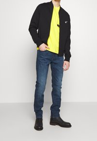 Černá bomber jacket na zip, žluté tričko s grafikou, modré džíny, černé kotníkové boty. Slim fit, hladká textura a minimalistický design.