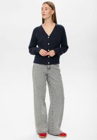 Mornarisk cardigan iz volnenega mešanega materiala z V-izrezom, šestimi belimi gumbi spredaj in rebrastimi manšetami, v kombinaciji s svetlo sivo širokima hlačami in rdečimi čevlji.