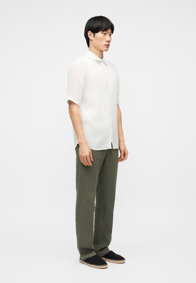 Άνδρας που στέκεται φορώντας λευκό πουκάμισο με κοντά μανίκια και κουμπιά, χακί παντελόνι και μαύρα παπούτσια slip-on, μπροστά από απλό φόντο.