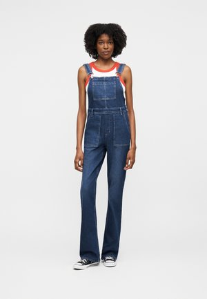 Femeie cu păr creț, purtând un top fără mâneci roșu și alb și o salopetă din denim albastru, cu adidași negri și albi, stând în picioare pe un fundal alb.