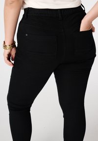 Persoon gekleed in zwarte, aansluitende broeken met achterzakken, met één hand in de zak en gouden armbanden om de linkerpols.