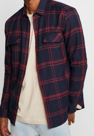 Man wearing an open navy and red plaid button-up shirt over a beige t-shirt and light blue jeans, standing against a plain background.