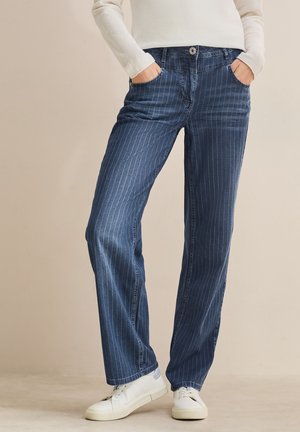 Blaue Denim-Jeans mit vertikalen weißen Streifen, ausgestattet mit einem Knopfverschluss, Fronttaschen und einem lockeren Schnitt. Getragen mit weißen Sneakers.
